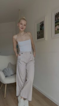 Calças de cintura alta em bege claro com bolsos laterais, design com pregas na frente, combinadas com uma t-shirt justa branca e ténis brancos.
