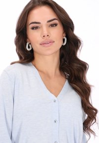 Világoskék kardigán puha anyagból, V-nyakú kivitelben és díszes gombokkal. Kerek, csillogó fülbevalóval viselve.