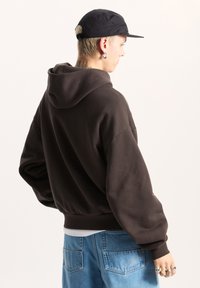Sudadera marrón con una capucha grande, bolsillo frontal y puños acanalados. Combinada con unos jeans de mezclilla azul claros y una gorra negra. Detalles de textura sutil.