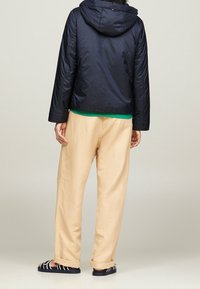 Personne portant un pantalon beige, une veste marine avec capuche, une chemise verte en dessous et des sandales noires avec des sangles blanches, se tenant de dos.