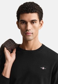 Čierny sveter s okrúhlym výstrihom, na hrudníku s malým vyšívaným logom. Model drží cez plece hnedú textúrovanú bundu.