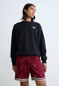 Schwarzer Sweatshirt mit kleinem weißen Logo und roten gemusterten Shorts mit weißen Streifen am Saum. Modell steht vor einem schlichten Hintergrund.