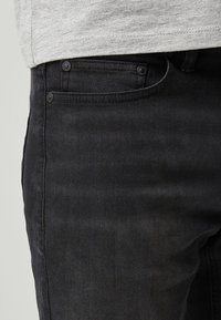 Mustad denim teksad, millel on kitsas lõige, tavaline vöökoht ja ees metallist riidest taskud. Kanga värv näib olevat pisut pleekinud.