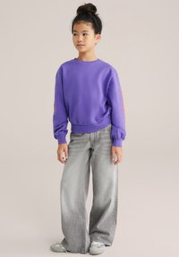 Pulover roșu-mov cu guler rotund și mâneci bufante, având un model detaliat portocaliu pe o mână. Asortat cu blugi lejeri, gri deschis și teniși.