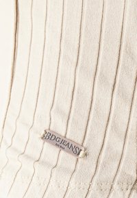 Beige ribbat tyg med en liten metalltagg där det står "BDG JEANS EST. 1998" sydd på materialet.