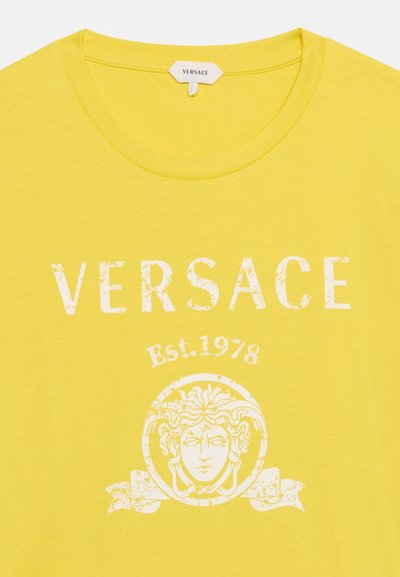 Κίτρινη μπλούζα Versace με λευκό τυπωμένο λογότυπο κεφαλής Μέδουσας, το κείμενο "VERSACE" πάνω και το "Est. 1978" κάτω από το όνομα της μάρκας.