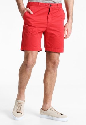 Homme portant un short rouge vif et des baskets beige, debout avec une main dans la poche, sur fond blanc.