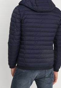 Marineblå isoleret pufferjakke med hætte, kviltet design og elastisk talje, parret med denimjeans.