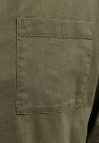 Cămașă verde măsliniu cu buzunar aplicat. Confecționată dintr-un material texturat cu margini cusute. Vedere în prim-plan care evidențiază textura și detaliile buzunarului.