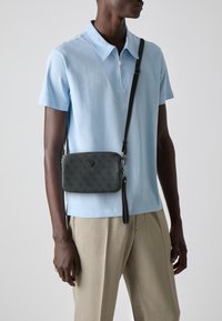 Bolso bandolera gris con un diseño en patrón y superficie texturizada, con cierre de cremallera y acento de marca, llevado sobre una camisa de cuello azul claro.