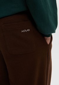 Brune sweatpants med en baglomme, der har et hvidt broderet logo. Stoffet ser blødt ud, og designet er afslappet pasform.