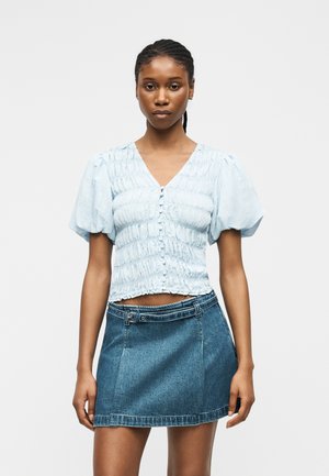 Kvinde med flettet hår iført lyseblå rynket bluse med knapper og pufærmer samt bælte i denim mininederdel, stående mod en ensfarvet baggrund.