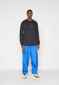 Schwarzes Langarmshirt mit Logodetail, kombiniert mit glänzenden blauen Hosen. Das Model trägt weiße und schwarze Sportschuhe.
