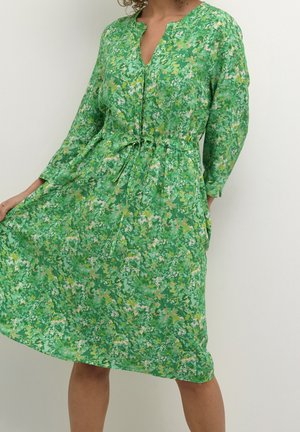 Vrouw draagt een groene bloemenprint jurk tot op de knie met lange mouwen, knoopjes aan de voorkant en een gebonden tailleband.