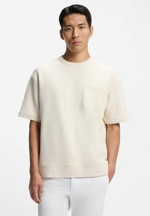 Junger Mann trägt ein übergroßes beige Kurzarmhemd mit Brusttasche und weiße Hose, steht vor einem einfarbigen weißen Hintergrund.