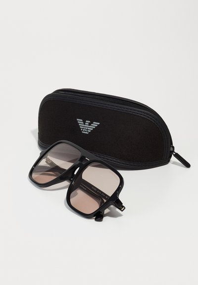 Gafas de sol negras de gran tamaño con lentes degradados, colocadas cerca de un estuche negro con cremallera que presenta un logo. Montura de plástico suave, forma cuadrada.