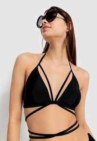 Zwarte bikini-top met driehoekige cups en kruislings gespen. Glad materiaal, minimalistisch ontwerp, gecombineerd met grote ronde zonnebrillen.