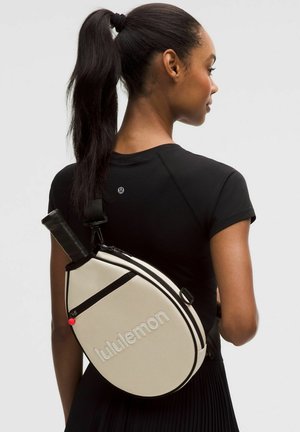 Frau mit Pferdeschwanz, die ein schwarzes Top trägt, trägt eine cremefarbene Lululemon Tennisschlägertasche mit schwarzen Akzenten auf dem Rücken.