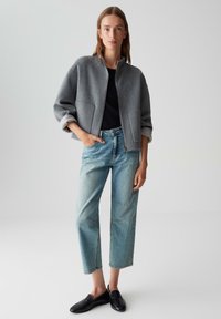 Vrouw in een grijze zip-up-jack, zwarte top, lichtblauwe cropped jeans en zwarte instapschoenen, staande met één hand in de zak.