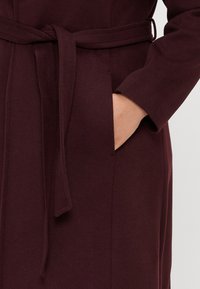 Παλτό μπορντό με ζώνη, κατασκευασμένο από υφασμάτινη υφή, διαθέτει πλαϊκή τσέπη και φαρδύ, ανοιχτό σχέδιο μανικιού.