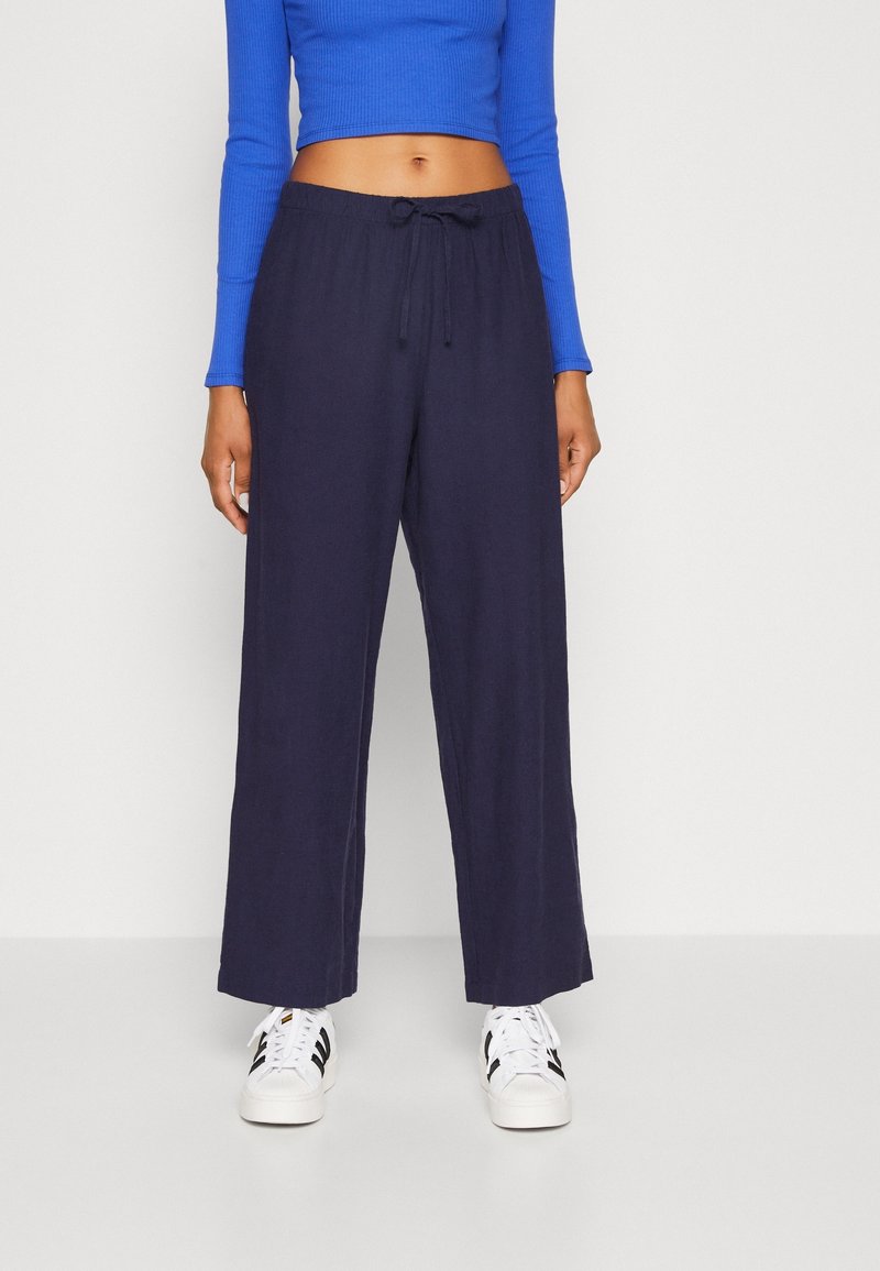 Pantalon large marine en tissu léger, doté d'une taille élastique et d'un cordon de serrage. Associé à des baskets blanches et un haut bleu.