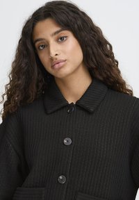 Jeune femme avec de longs cheveux bouclés et foncés, portant une veste noire côtelée avec de grands boutons noirs et un col pointu, sur un fond blanc.