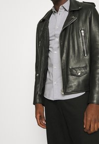 Giacca da biker in pelle nera con cerniere e bottoni in argento, caratterizzata da un taglio corto, tasche laterali e una texture liscia. Indossata sopra una camicia grigia chiara.