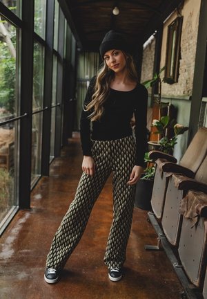 Zwarte langwerpige top gecombineerd met hoog-wijze, gepatreerde wijde broek in donkergroen en crème, gedragen met zwarte sneakers.