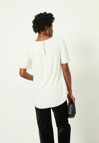 Weiße Kurzarmbluse aus strukturiertem Stoff, mit Knopfdetail auf der Rückseite und lockerer Passform. Kombiniert mit schwarzen Hosen und einer gehaltenen Tasche.