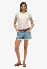 Camiseta de manga corta, de color gris claro, con mangas raglán en azul claro y logotipo bordado; combinada con pantalones cortos de mezclilla y un cinturón tejido marrón con sandalias.
