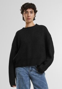 Zwarte gebreide trui met een ribbelstructuur, brede mouwen en een ronde halslijn, gedragen over blauwe jeans. Casual, ontspannen pasvorm.