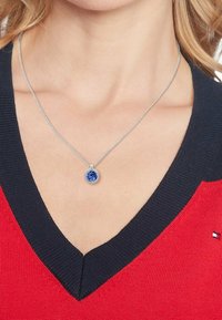 Collier en chaîne argentée avec un pendentif en pierre bleue ronde, présentant une surface texturée, porté sur un pull rouge et marine à col en V.