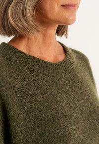 Groene wollen trui met een zachte, textuurafwerking, voorzien van een ronde halslijn en ribbelranden. Close-up van de halslijn en schouderpartijen.