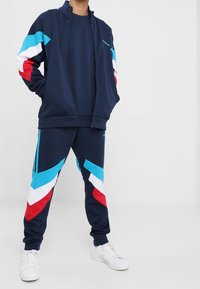 Marinträningsoverall med geometriska färgaccenter i blått, rött och vitt. Består av en zip-up jacka och avsmalnande byxor. Tillverkad av mjukt, andningsbart tyg.