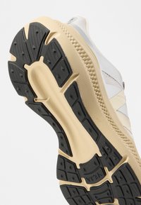 Πάτος λευκού και μπεζ αθλητικού παπουτσιού με μαύρη, υφή πέλματος σχεδιασμένη για κράτημα και ανθεκτικότητα.