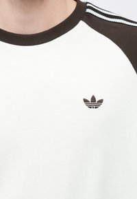 Λευκό φούτερ από βαμβακερό μείγμα με στρογγυλή λαιμόκοψη και καφέ ραγκλάν μανίκια, με μικρό καφέ λογότυπο Adidas στο αριστερό στήθος και ριγωτές λεπτομέρειες.