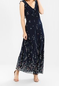 Maxi-jurk van marineblauwe stof met een bloemenpatroon in roze en groen. V-hals, ruches bij de schouder en een vloeiende rok.