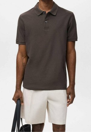 Mann trägt dunkelbraunes kurzärmeliges Poloshirt und hellbeige Shorts, hält eine schwarze Tragetasche vor schlichter Hintergrund.