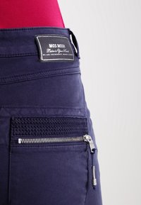 Marineblauwe broek met een zwart label waarop "MOS MOSH" staat. Beschikt over een textuurzak met een ritsluiting en contrasterende stiksels.
