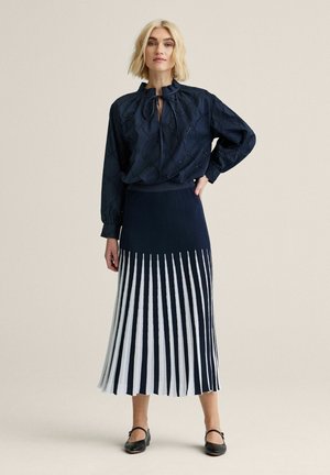 Lyshåret kvinde står iført en navyblå bluse med hulmønster og bindebånd ved halsen samt en navyblå plisseret nederdel med hvide lodrette striber, sorte Mary Jane-sko.