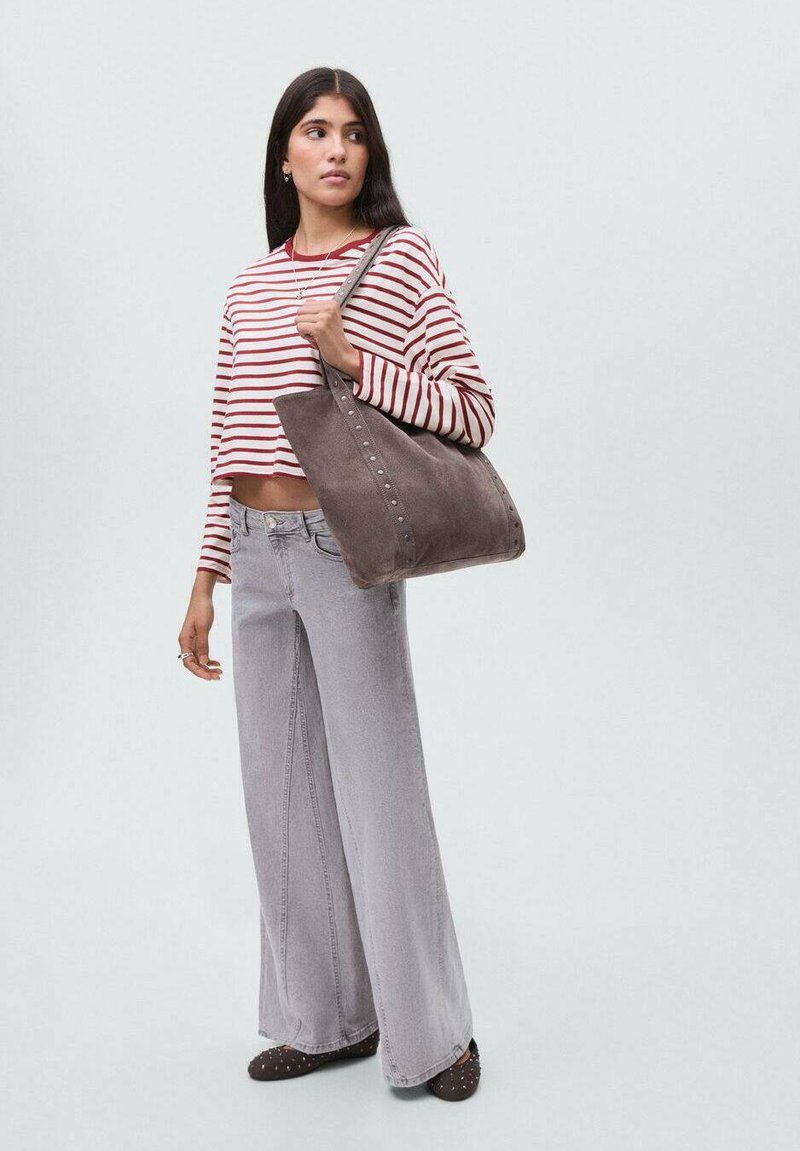 Kvinna iklädd en röd och vit randig långärmad crop top, vida grå jeans, bär en stor grå mocka väska över axeln.
