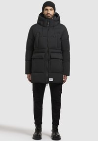 Black puffer jacket with a zip closure, hood, and two large front pockets. Smooth texture, stitched design, worn with black cargo pants and boots.