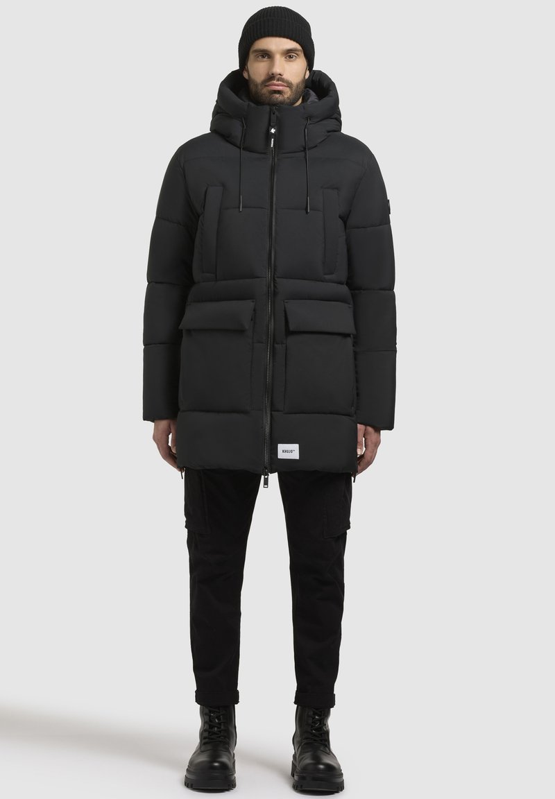Black puffer jacket with a zip closure, hood, and two large front pockets. Smooth texture, stitched design, worn with black cargo pants and boots.