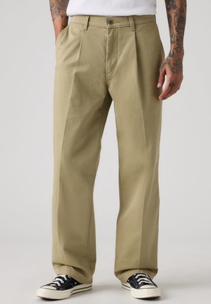 Mand iført beige plisserede bukser og sorte sneakers med hvide såler, stående med hænderne nær lommerne mod en ensfarvet baggrund.