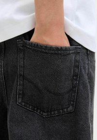 Main dans la poche arrière d'un jean en denim noir porté avec une chemise blanche.