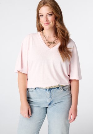 Jeune femme aux longs cheveux roux portant un chemisier rose clair à col en V, des colliers dorés superposés, un jean bleu clair et une ceinture beige, debout devant un fond blanc.