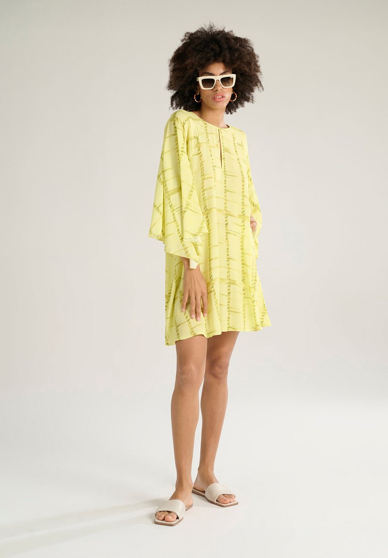 Abito giallo con una vestibilità morbida, caratterizzato da maniche a campana e un design a fantasia. Abbinato a sandali scivolosi neutri e occhiali da sole oversize.