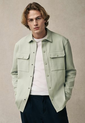 Light green button-up shirt in a soft material, featuring two front chest pockets and a classic collar, worn over a white t-shirt.