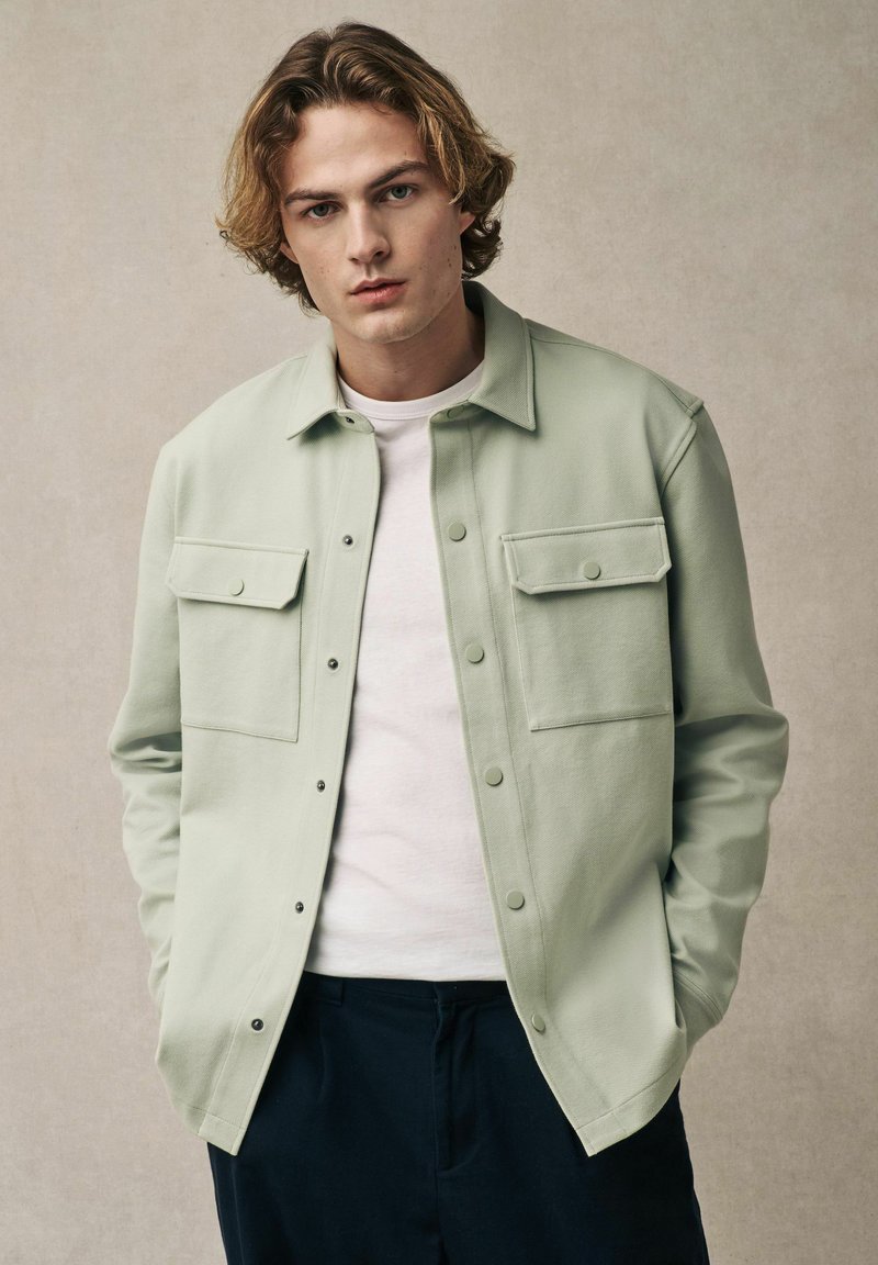 Camicia verde chiaro con bottoni realizzata in un materiale morbido, dotata di due tasche frontali sul petto e colletto classico, indossata sopra una t-shirt bianca.