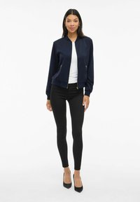 Kvinna står upp, iförd en marinblå bomberjacka över vit topp, svarta skinny jeans och svarta högklackade skor framför en vit bakgrund.